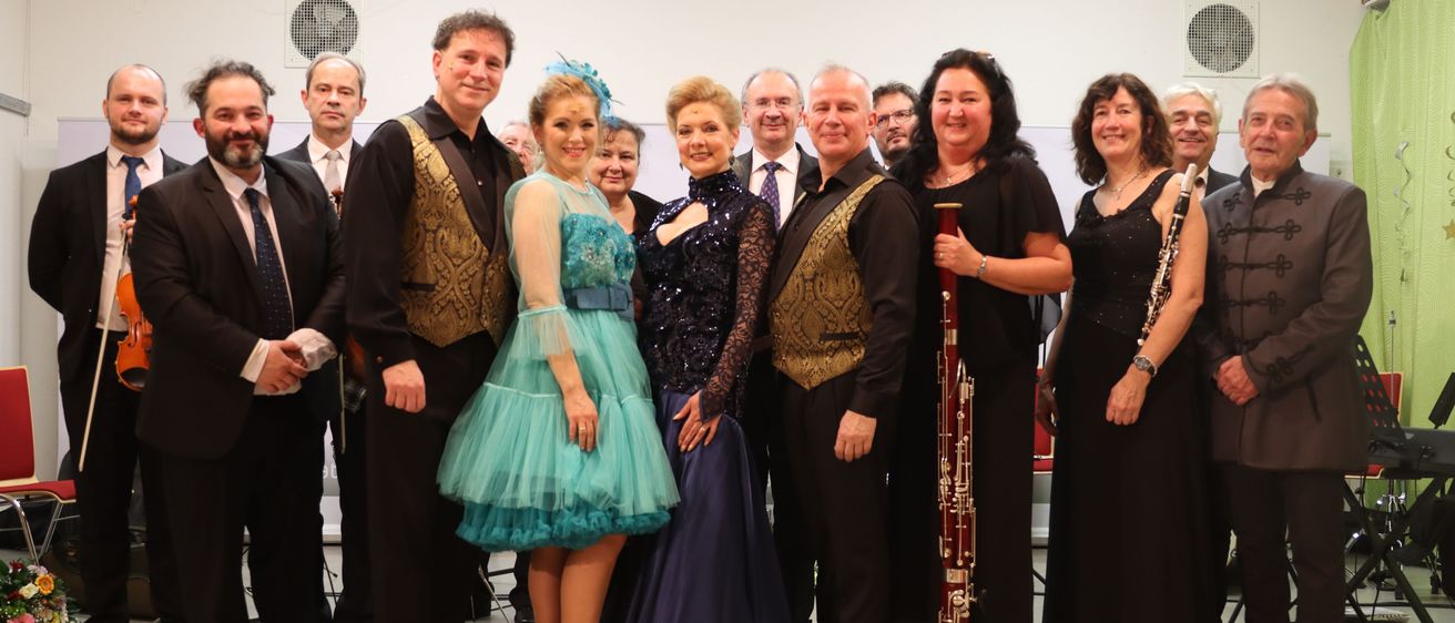 Eine Gruppe von Musikern posiert für ein Foto. Die Gruppe besteht aus einer Frau in einem türkisfarbenen Kleid, einer Frau in einem blauen Kleid und einem Mann, der ein Fagott hält. Die Männer und Frauen tragen formelle Kleidung.