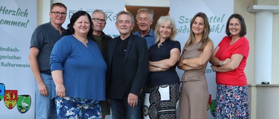 A group of people, including men and women, are standing together in front of a white banner with green text. They are smiling and posing for a photo.