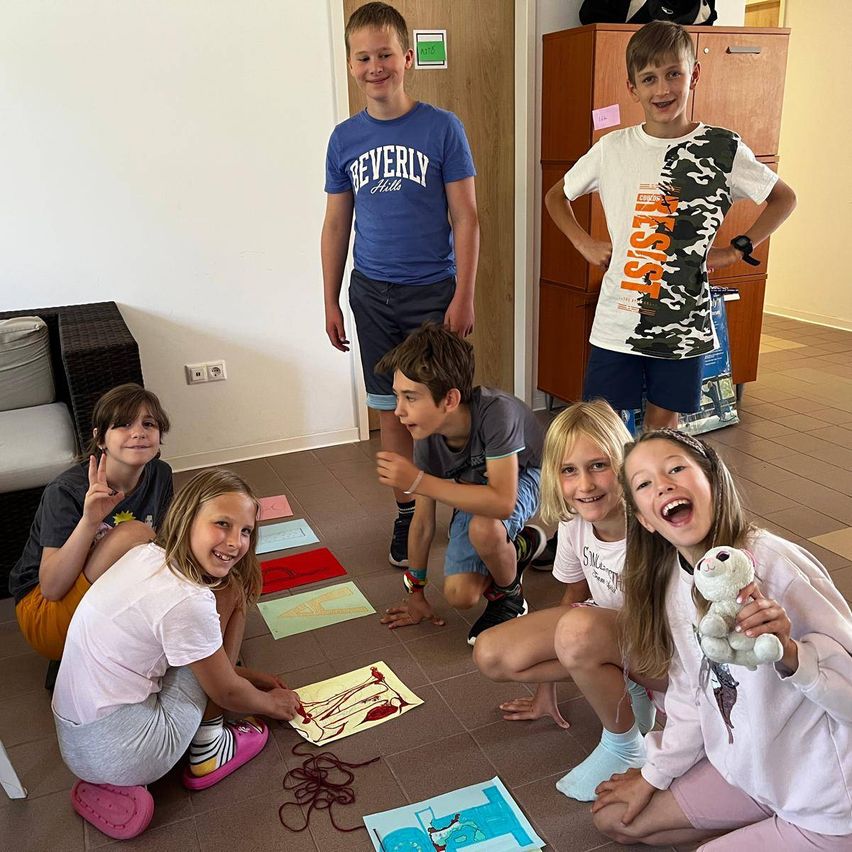 Gruppe von Kindern sitzt auf dem Boden und macht ein Kunstprojekt. Der Boden ist mit Papieren und Garnstücken bedeckt.
