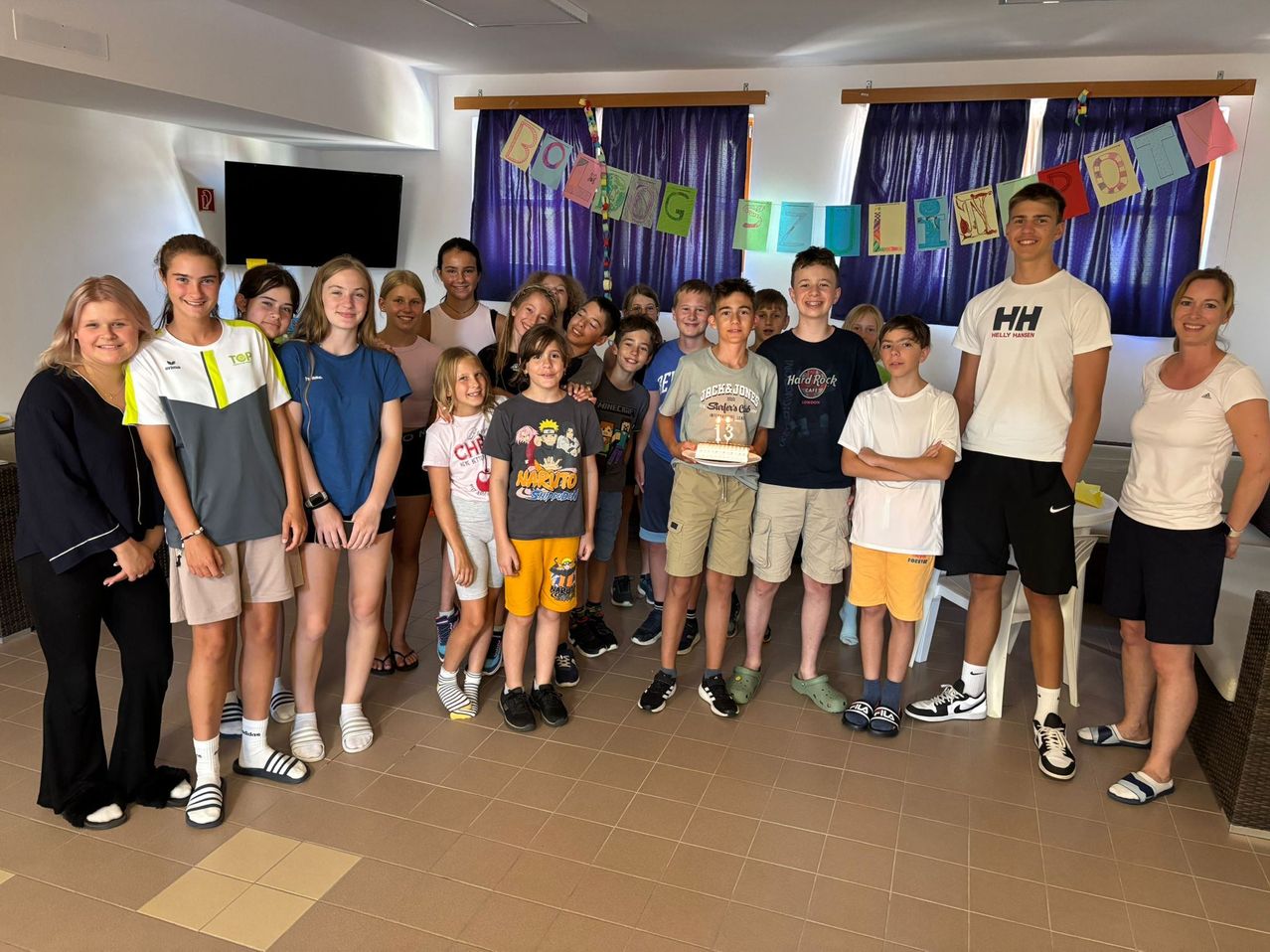 Eine Gruppe von Kindern und Erwachsenen steht in einem Raum und posiert für ein Foto. Die Kinder tragen Freizeitkleidung, und zwei von ihnen tragen Turnschuhe. Eine Person in der Mitte hält einen Kuchen, und neben ihm steht eine Person. Hinter ihnen befinden sich Vorhänge, ein Fernseher und ein weißer Tisch.
