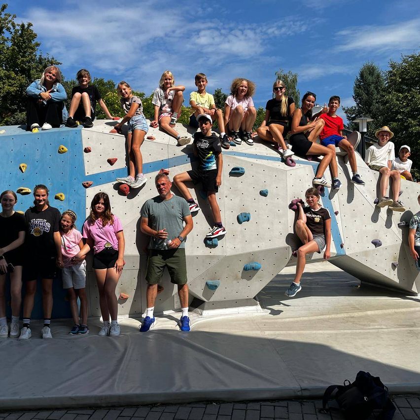 Eine Gruppe von Menschen, darunter Erwachsene und Kinder, posiert für ein Foto an einer großen Kletterwand an einem sonnigen Tag.