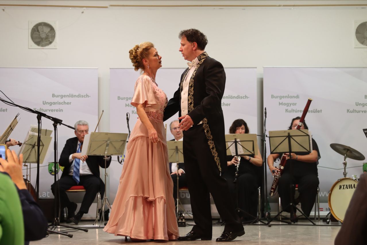 Ein Paar in formeller Kleidung tanzt auf der Bühne, während eine Band im Hintergrund spielt. Die Frau trägt ein rosa Kleid und der Mann einen schwarzen Anzug. Sie sehen sich mit konzentrierten Gesichtsausdrücken an. Die Band besteht aus einem Klarinettisten und einem Fagottspieler. Der Hintergrund zeigt weiße Banner mit fremdsprachigem Text.