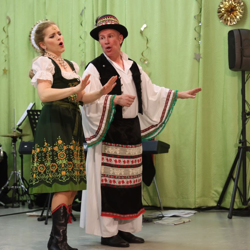 Ein Mann und eine Frau treten auf der Bühne in traditioneller Volkstracht auf. Der Mann trägt einen Hut und eine Weste, während die Frau eine Halskette trägt. Sie beide gestikulieren. Hinter ihnen befindet sich ein grüner Vorhang, ein Lautsprecher und Musikinstrumente.