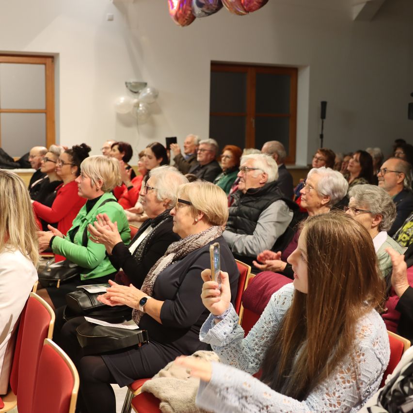 Ein Raum voller Publikum, einige klatschen und andere schauen auf ihre Handys. Einige tragen Brillen, Schals und Jacken. Der Raum hat weiße Wände, Glasfenster und Ballons.