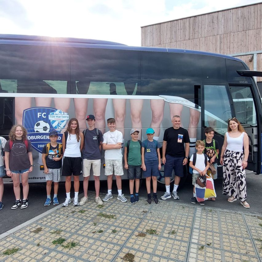 Eine Gruppe von Kindern und Erwachsenen steht vor einem Bus mit einem Fußballteam-Logo an der Seite.