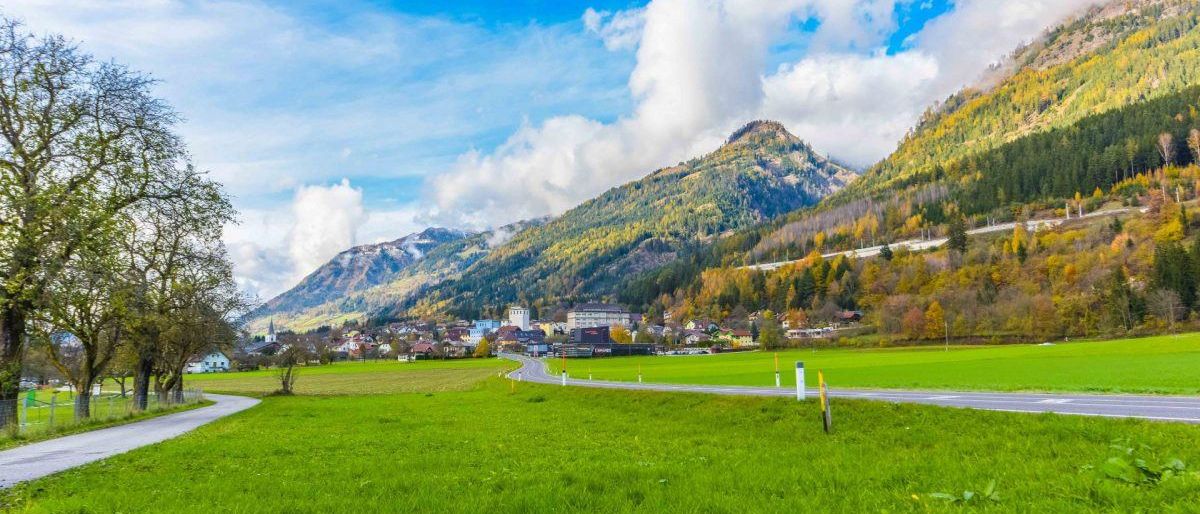 Eine friedliche Landschaft mit einem saftig grünen Feld, einer kurvenreichen Straße und von Bäumen bedeckten Bergen. Der Himmel ist klar mit flauschigen Wolken.