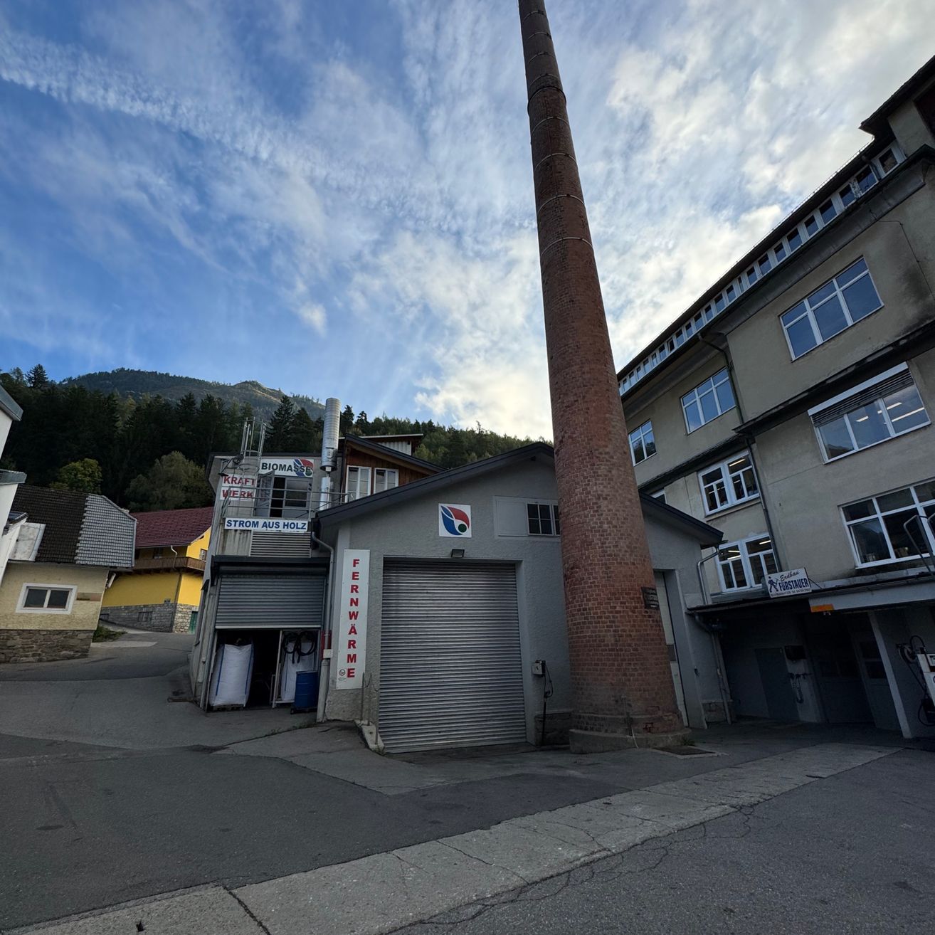 Ein Industriegebiet mit einem hohen Schornstein und einem Gebäude mit der Aufschrift Fernwarm. Ein Schild steht für Strom aus Holz, und der Himmel ist blau mit Wolken.