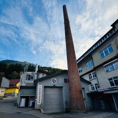 Ein hoher Schornstein steht neben einem Gebäude mit einer großen Garagentür, einem Logo und einem Schild, das Fernwärme liest. Ein blauer Himmel mit verstreuten Wolken ist im Hintergrund.