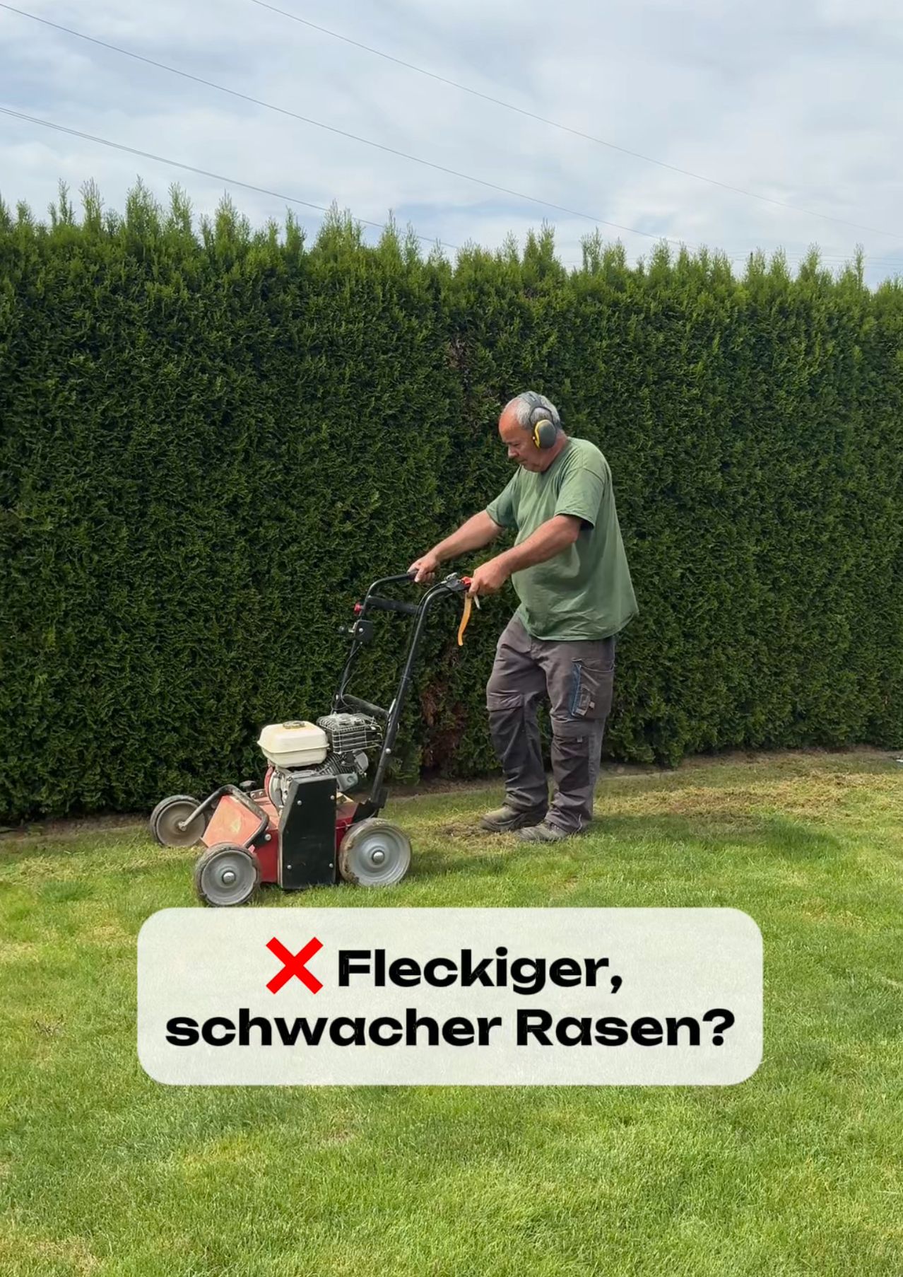 Ein Mann in einem grünen Hemd und grauen Hosen steht neben einer Hecke und benutzt einen roten Rasenmäher mit gelbem Griff.