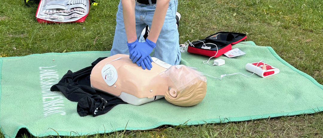 Eine Person übt CPR an einem Mannequin auf einer grünen Decke in einem Grasbereich mit medizinischen Geräten in der Nähe.