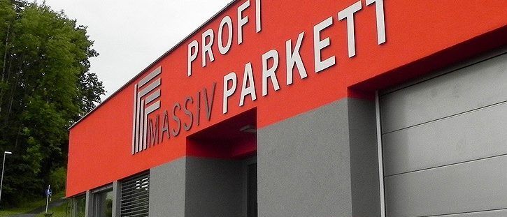 Ein orange-graues Gebäude mit der Aufschrift 'Profi Massiv Parkett' steht draußen. Das Gebäude hat zwei Garagentore, zwei Fenster und einen Rasen.