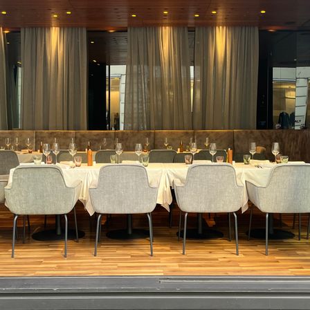 Ein elegantes Restaurant mit einem langen Tisch für das Essen. Die Stühle sind um den Tisch herum angeordnet, und es gibt Weingläser und andere Tischwaren. Der Raum ist mit Vorhängen dekoriert.