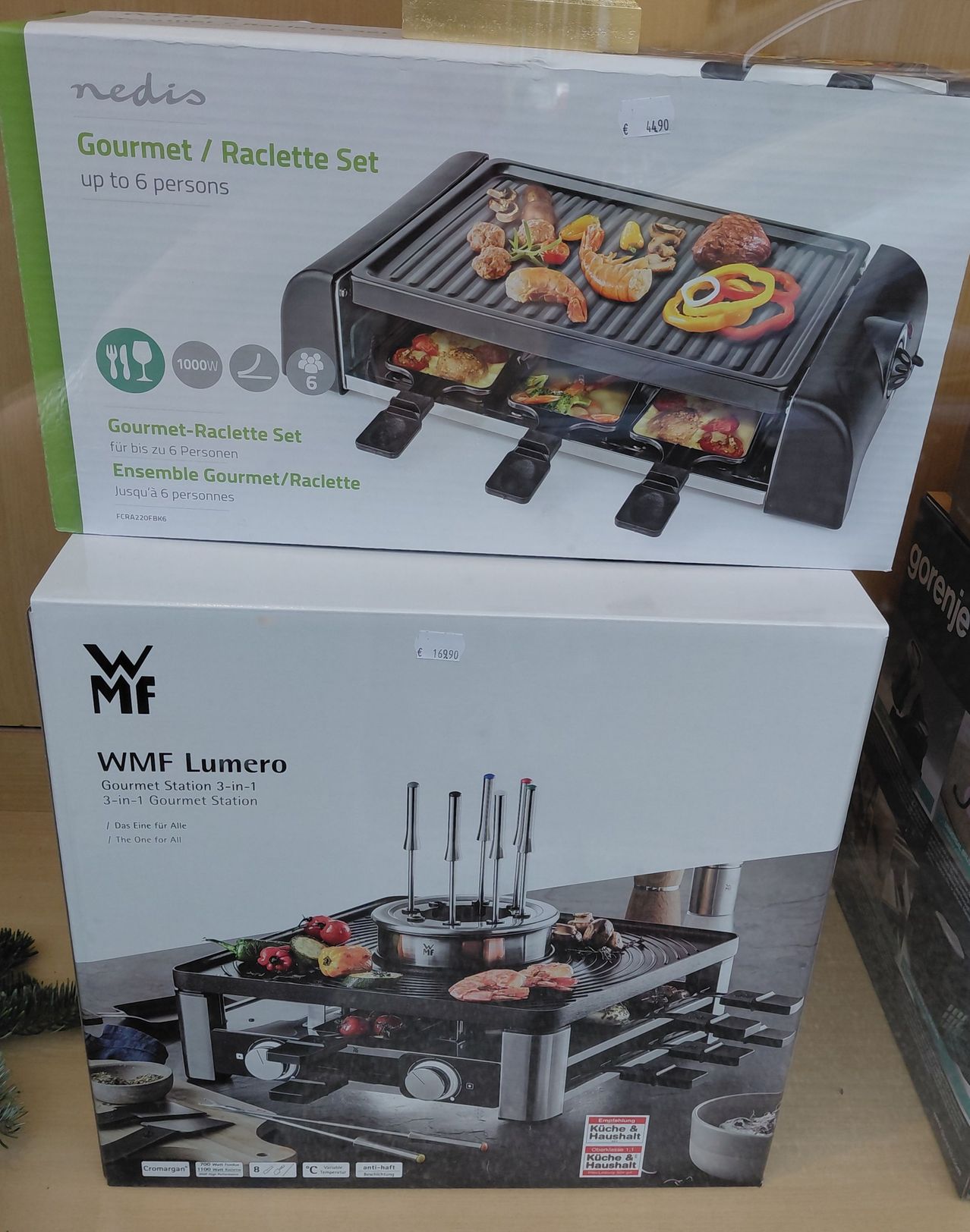 Zwei Kartons einer WMF Lumero Gourmet Station 3-in-1 sind gestapelt. Sie ist für bis zu sechs Personen ausgelegt und bietet Grill-, Fondue- und Raclette-Optionen. Der obere Karton zeigt die Funktionen und Kapazität des Produkts.
