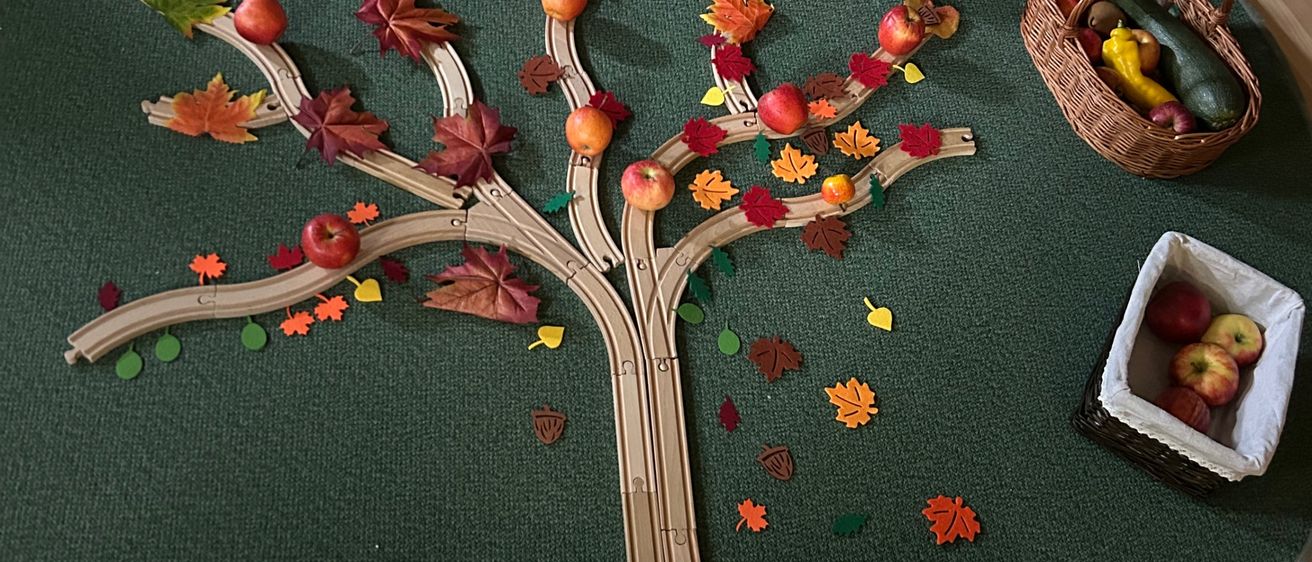 Ein grüner runder Teppich mit einem hölzernen Apfelbaum, umgeben von verschiedenen Früchten und Gemüse. Ein Korb mit Äpfeln und ein Korb mit Obst und Gemüse sind in der Nähe.