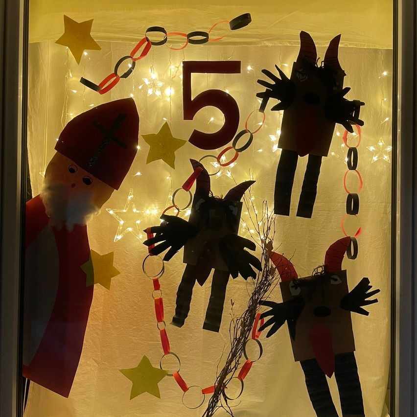 Eine festliche Dekoration zeigt eine rote Weihnachtsmann-Figur, eine rote Nummer 5 und drei handgefertigte Papierfiguren. Die Szene umfasst gelbe Sterne und rote Papierketten, alles beleuchtet von warmen Lichtern.