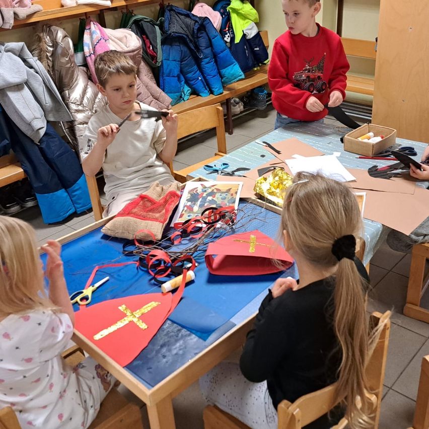 Mehrere Kinder sitzen um einen Tisch herum und beschäftigen sich mit Bastelarbeiten. Sie haben Materialien wie Scheren, Bänder und rotes Papier vor sich. Ein Junge in einem roten Sweatshirt steht in der Nähe.