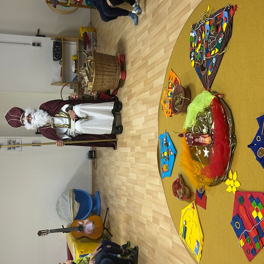 Ein festliches Ereignis mit einer Person in Santa Claus-Verkleidung. Ein runder Tisch ist mit verschiedenen Dekorationen geschmückt. Eine Gitarre und ein Korb stehen in der Nähe.