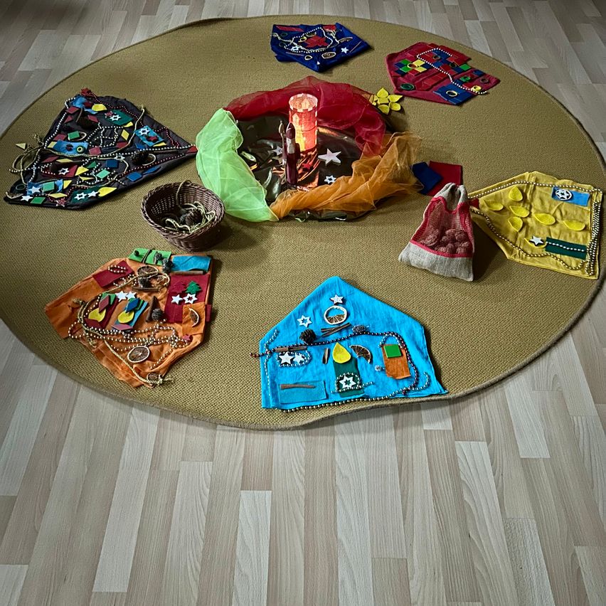 Ein runder Teppich mit einer Kerze und bunten Gegenständen darauf. Es gibt Taschen und Körbe mit verschiedenen Objekten darum herum. Der Teppich liegt auf einem Holzboden.