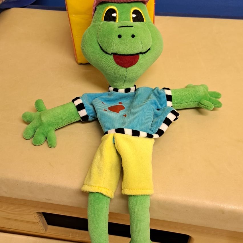 Ein grüner Plüschfrosch sitzt auf einer beigen Oberfläche und trägt ein blaues Shirt mit einem roten Herzen und gelben Shorts. Der Frosch hat einen Hut und ausdrucksstarke Augen.