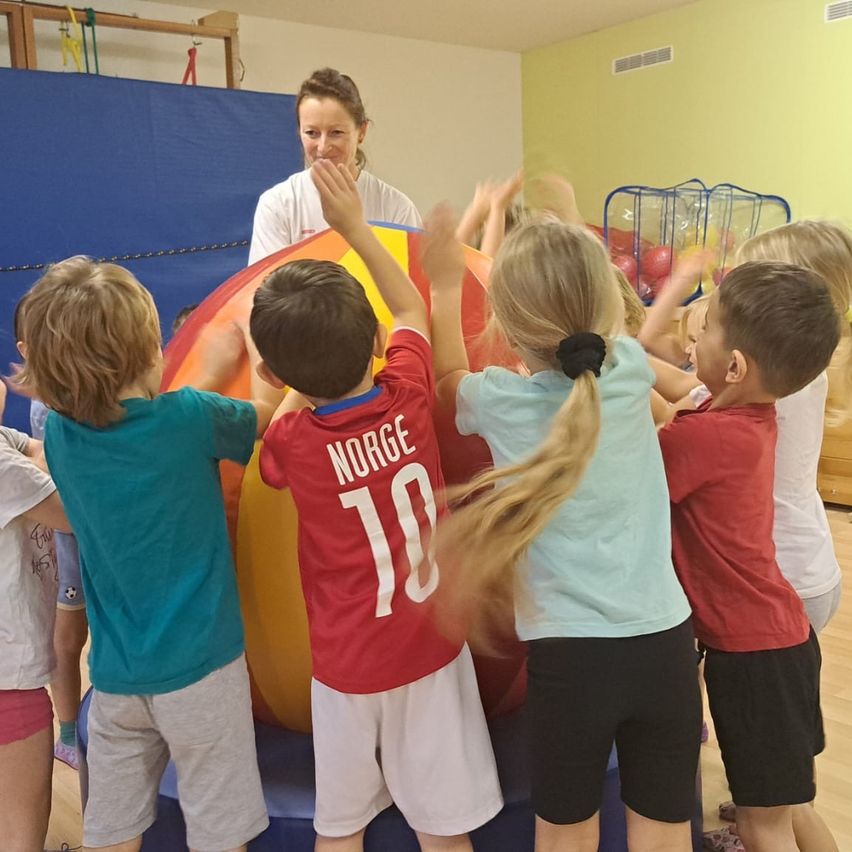 Mehrere Kinder versammeln sich um einen großen aufblasbaren Ball und tragen verschiedene farbige T-Shirts. Eine Frau steht hinter ihnen, lächelt und beaufsichtigt die Aktivität.