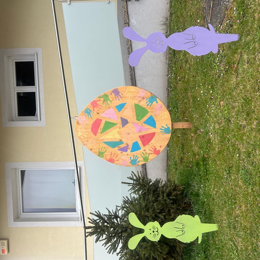 Ein Osterei mit bunten Handabdrücken steht in einem Grasbereich, neben zwei lila und grünen Hasenausschnitten. Ein Gebäude mit zwei Fenstern befindet sich dahinter.