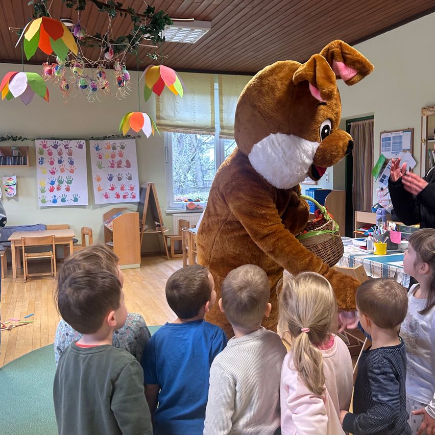 In einem Kindergartenraum sind Kinder um einen Maskottchen versammelt, das als Hund verkleidet ist. Bunte Dekorationen und Handabdrücke sind an den Wänden, und es gibt Tische mit Spielzeug.