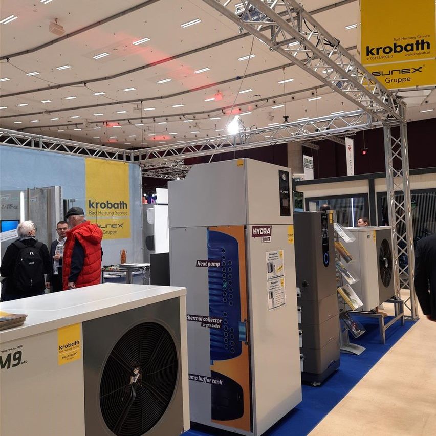 An indoor exhibition with various booths, one labeled Krobath. People are gathered around a blue carpet. Some booths have air conditioning units and banners.