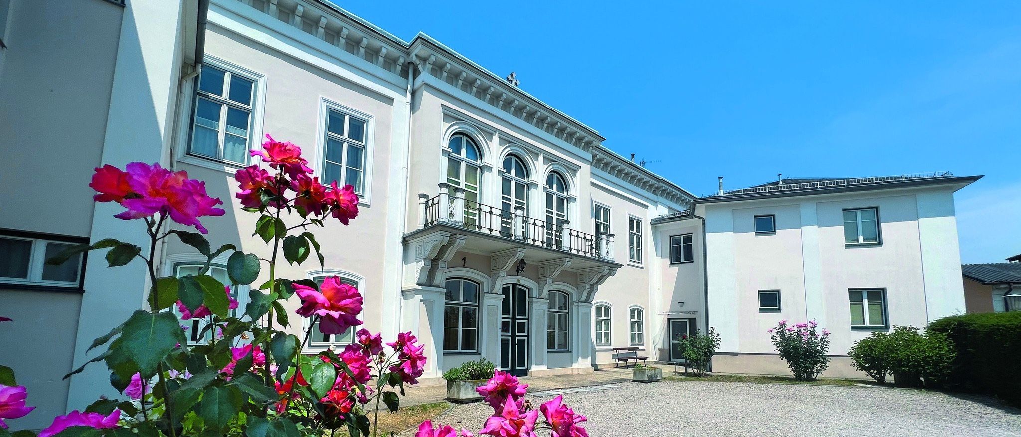 Ein weißes Gebäude mit Bogenfenstern, Balkonen und einer Kiesauffahrt, geschmückt mit rosa Rosen vor dem Gebäude.