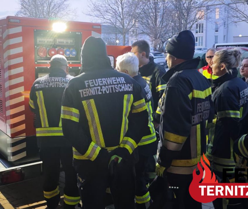 Eine Gruppe von Feuerwehrleuten aus Ternitz-Pottschach steht vor einem roten Feuerwehrauto. Sie tragen Uniformen mit reflektierenden Streifen und Mützen. Ein rotes Feuerlogo ist im unteren rechten Bildbereich sichtbar.