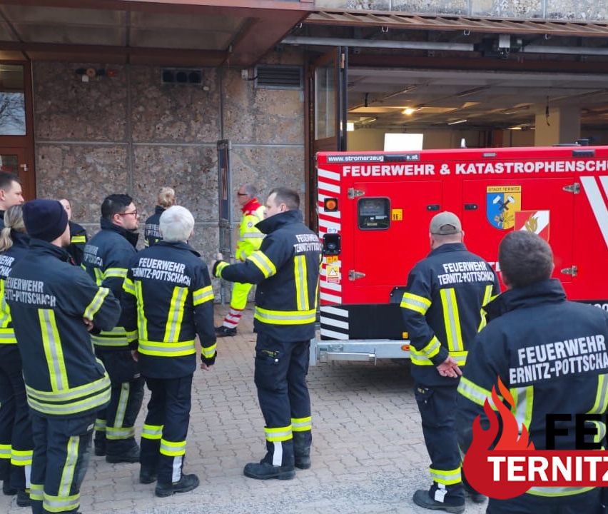 Feuerwehrleute in blauen Uniformen stehen vor einem Feuerwehrhaus. Ein Mann spricht mit anderen. Ein rotes Feuerwehrauto ist in der Nähe geparkt.