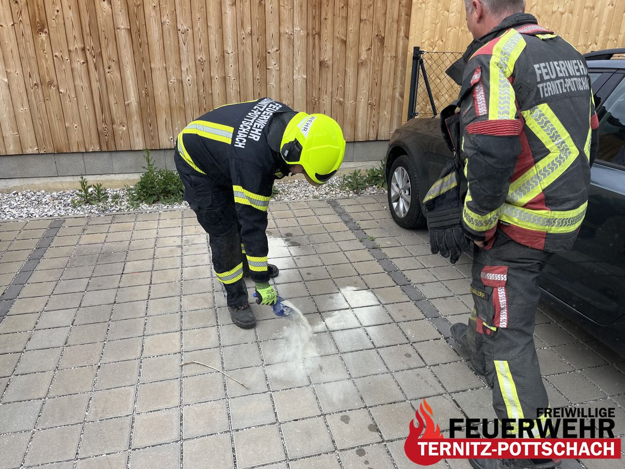 A firefighter is pouring a substance on the ground. Another firefighter stands beside him, both wearing yellow helmets. A black car is parked behind them.