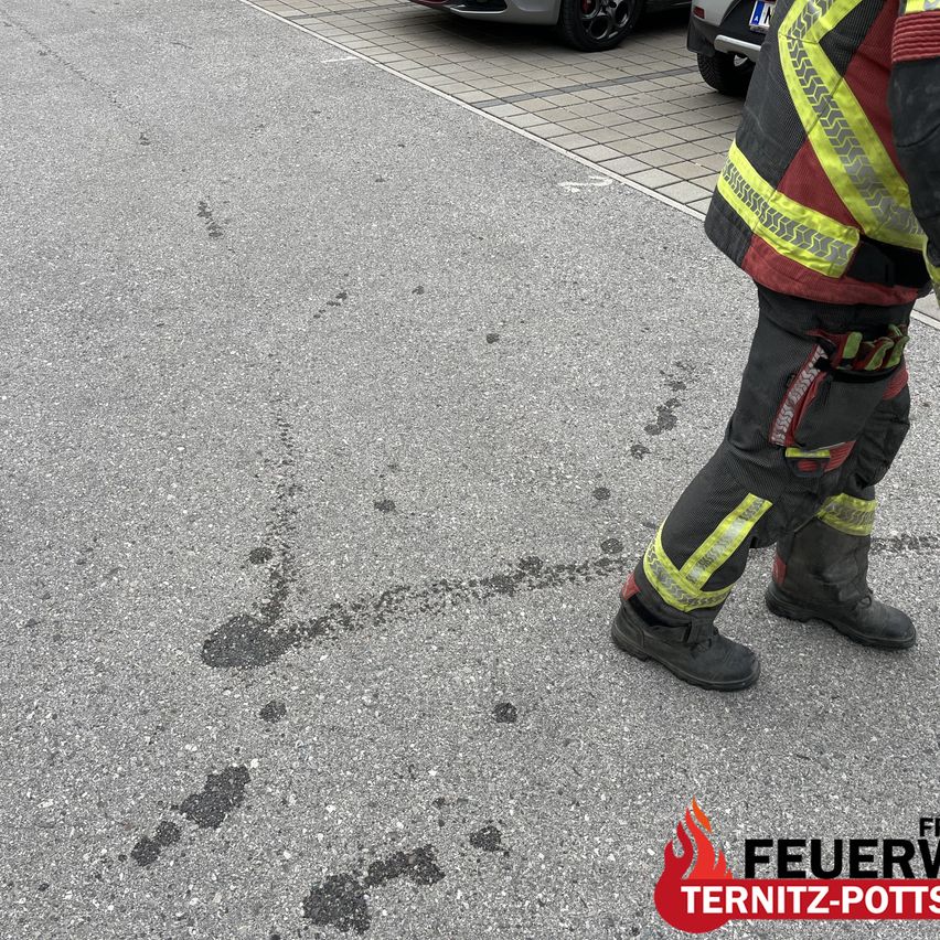 A firefighter in a reflective vest walks on a wet road with oil stains. A car with a license plate is parked nearby.