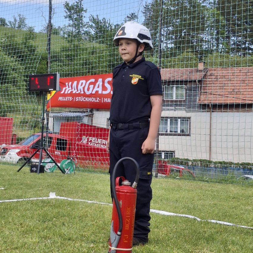 Ein Kind in Feuerwehruniform steht neben einem Feuerlöscher auf einem grünen Feld mit einem roten Banner im Hintergrund.