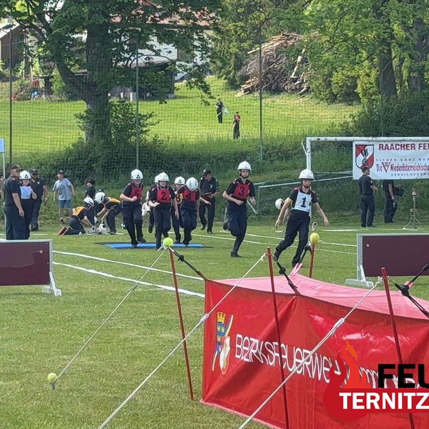 Eine Gruppe von Personen in Helmen ist an einem Sportevent auf einem Feld beteiligt. Sie rennen in Richtung eines Tores und scheinen ein Spiel zu spielen. Ein roter Banner mit dem Text 'BERZSFUERWE' ist vor ihnen platziert.