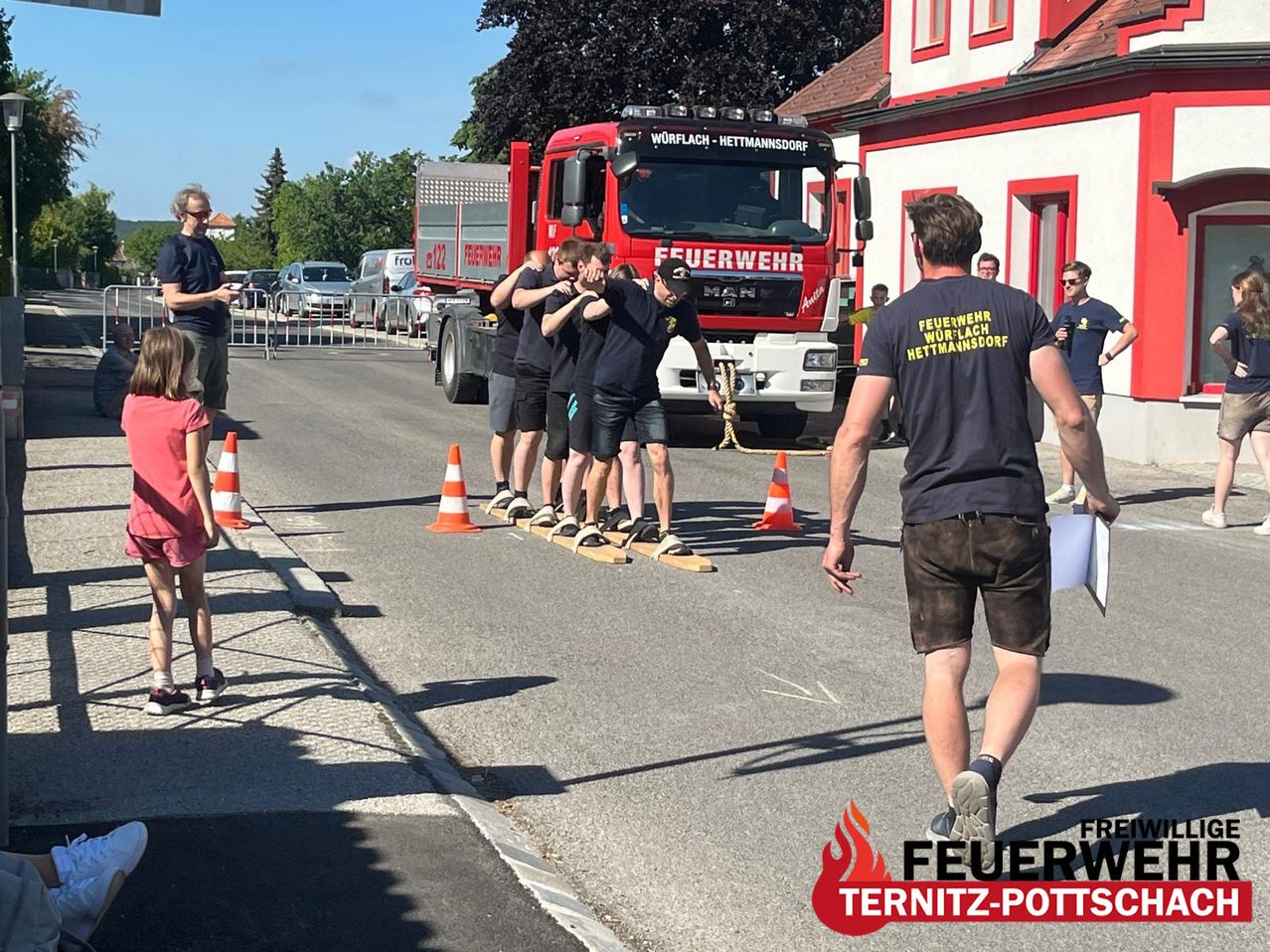 Eine Gruppe Feuerwehrleute übt das Seilklettern auf einer Straße in der Nähe eines Feuerwehrwagens. Zuschauer beobachten und ein Mann geht vorbei.