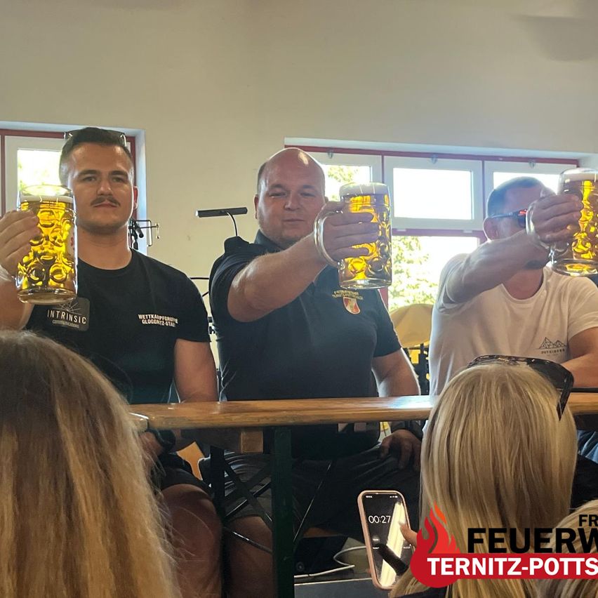 Vier Männer sitzen um einen Tisch herum, jeder hält ein großes Bierglas, wahrscheinlich im Feiern. Eine Frau mit langen Haaren sitzt hinter ihnen. Ein Wasserzeichen sagt 'Feuerwehr Ternz-Potts'.