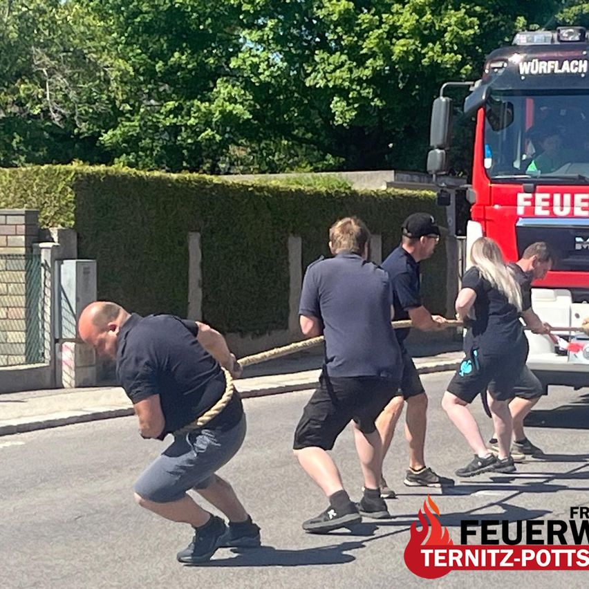 Fünf Feuerwehrleute ziehen an einem sonnigen Tag an einem Seil. Sie tragen dunkle Uniformen und Turnschuhe. Der LKW ist mit dem Wort "Feuer" auf der Straße geparkt.