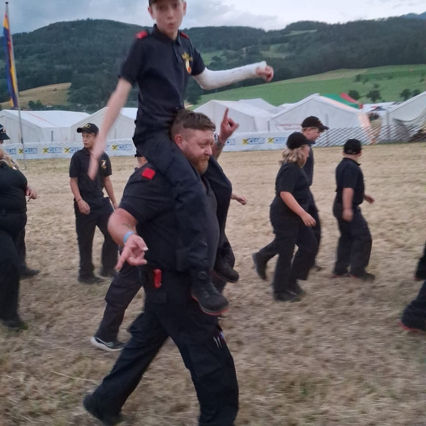 Ein Junge in Uniform reitet auf den Schultern eines Mannes in einem Feld, während andere in Uniform hinter ihnen stehen.