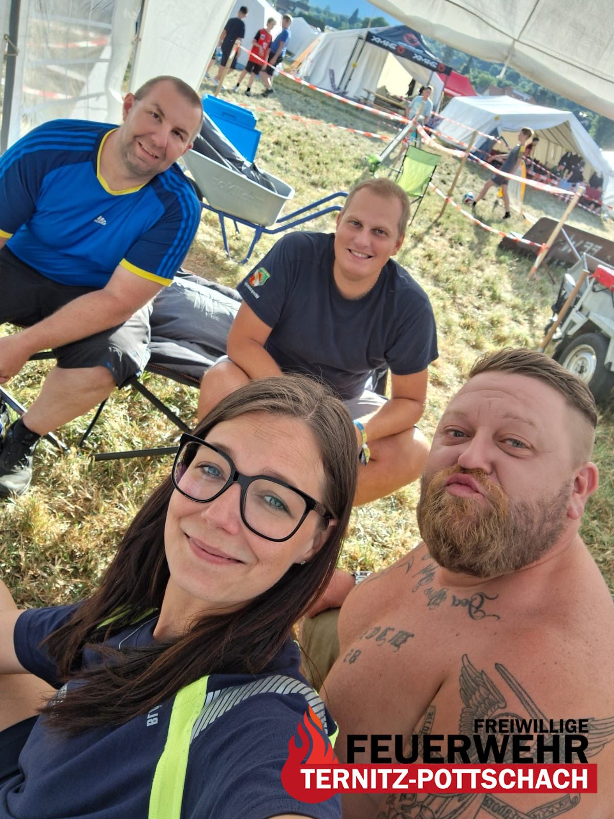 Vier Menschen lächeln und posieren für ein Foto in einem Grasbereich. Zwei Männer sitzen auf Stühlen und eine Frau sitzt auf dem Boden. Sie tragen alle Freizeitkleidung. Hinter ihnen stehen Zelte, ein Schubkarren und ein Fußball.