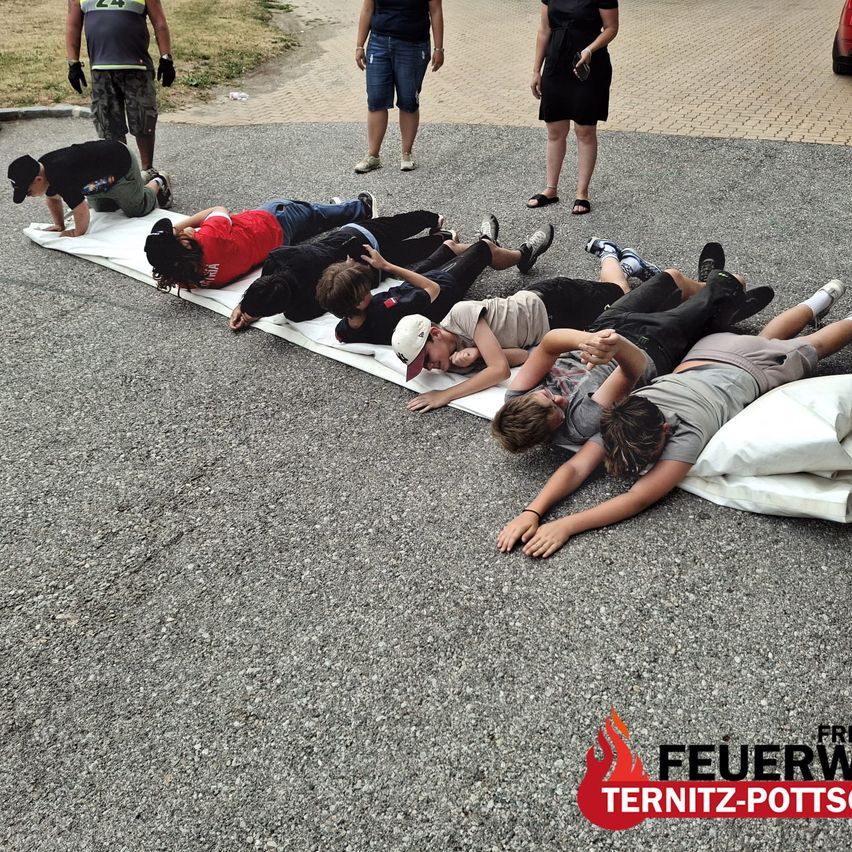 Eine Gruppe von Menschen liegt auf einem langen weißen Laken auf dem Boden, möglicherweise an einer Protestaktion beteiligt. Einige Leute stehen um sie herum, und einer hält eine Kamera. Das Bild hat einen Wasserzeichen mit dem Text 'FEUERW TERNITZ-POTTS'.