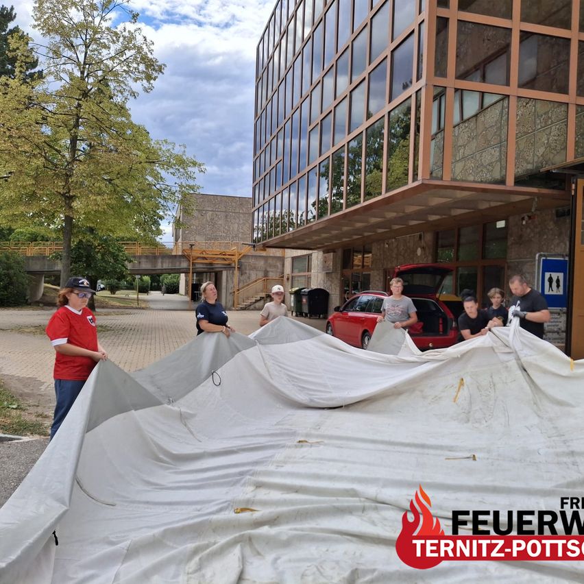 Eine Gruppe von Menschen richtet eine große weiße Plane vor einem Gebäude mit Glasfenstern auf. Ein roter Wagen mit offenem Kofferraum ist in der Nähe geparkt.