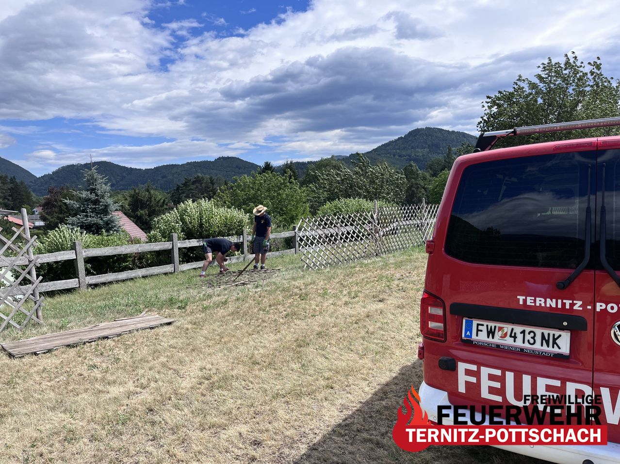 Zwei Personen arbeiten an einem Zaun in einem Grasfeld, mit einem roten Feuerwehrwagen im Hintergrund. Der Himmel ist bewölkt.