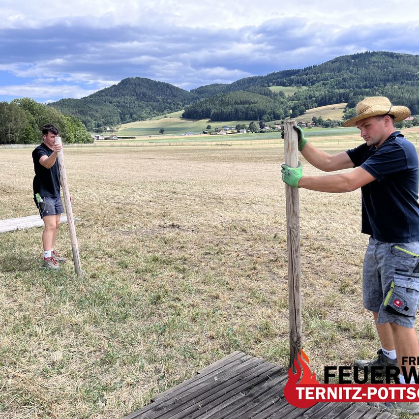 Zwei Männer installieren einen Holzpfosten in einem Feld mit Bergen im Hintergrund. Ein Mann trägt einen Strohhut und Handschuhe.