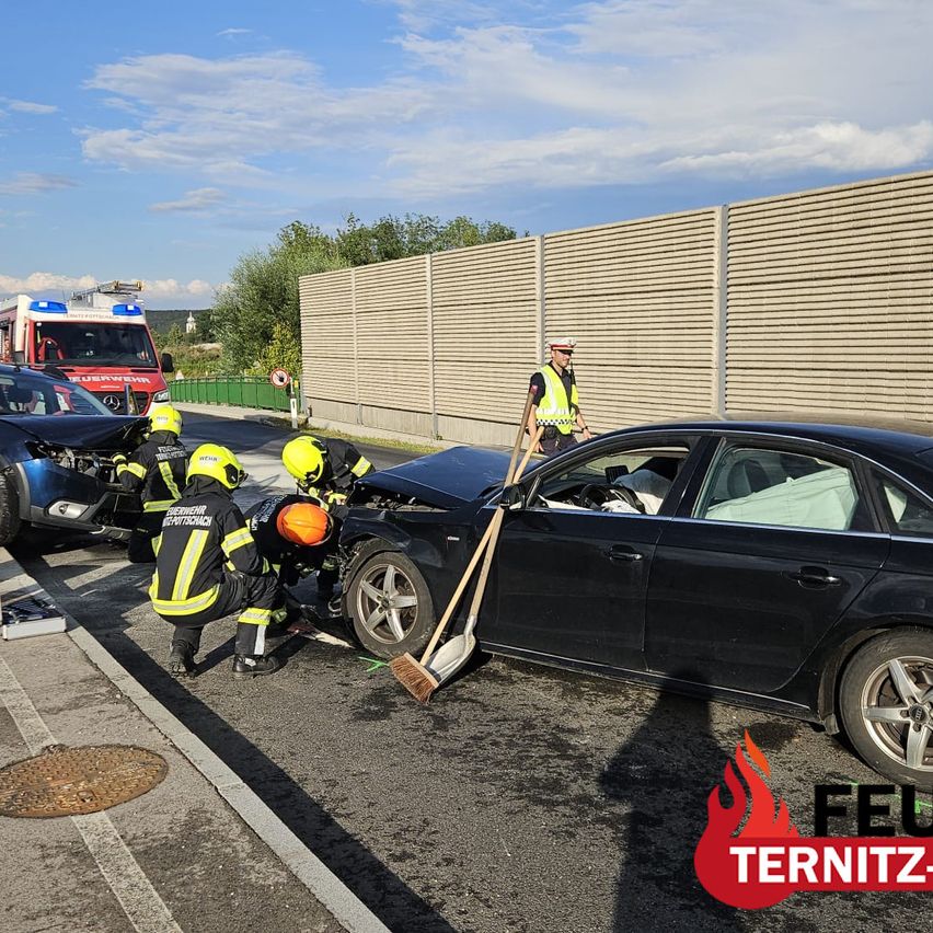 Firefighters in yellow uniforms are working on a damaged black car on the side of the road. A fire truck is parked behind them.