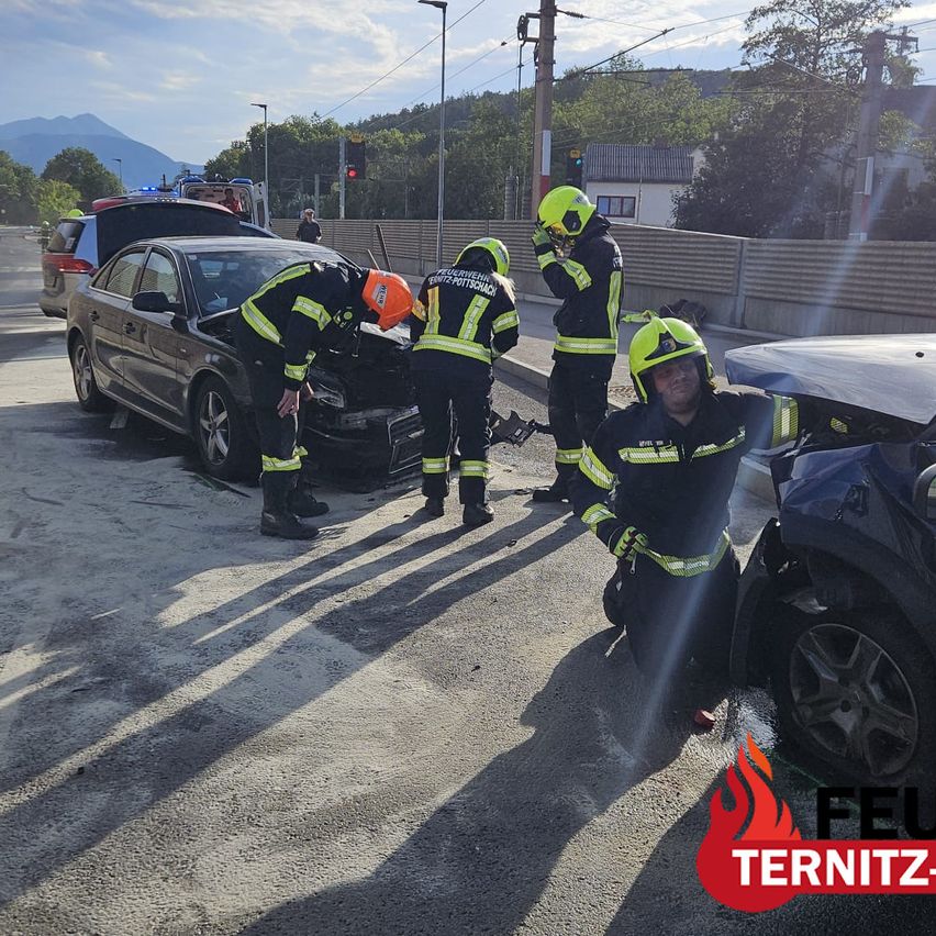 Firefighters in protective gear are attending to a car accident on a road, with one person examining the vehicle and another standing nearby.
