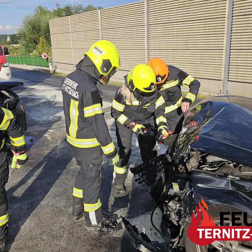 Three firefighters in protective gear are inspecting a damaged car on the side of the road. One is holding a tool, and the car has visible damage to its front.