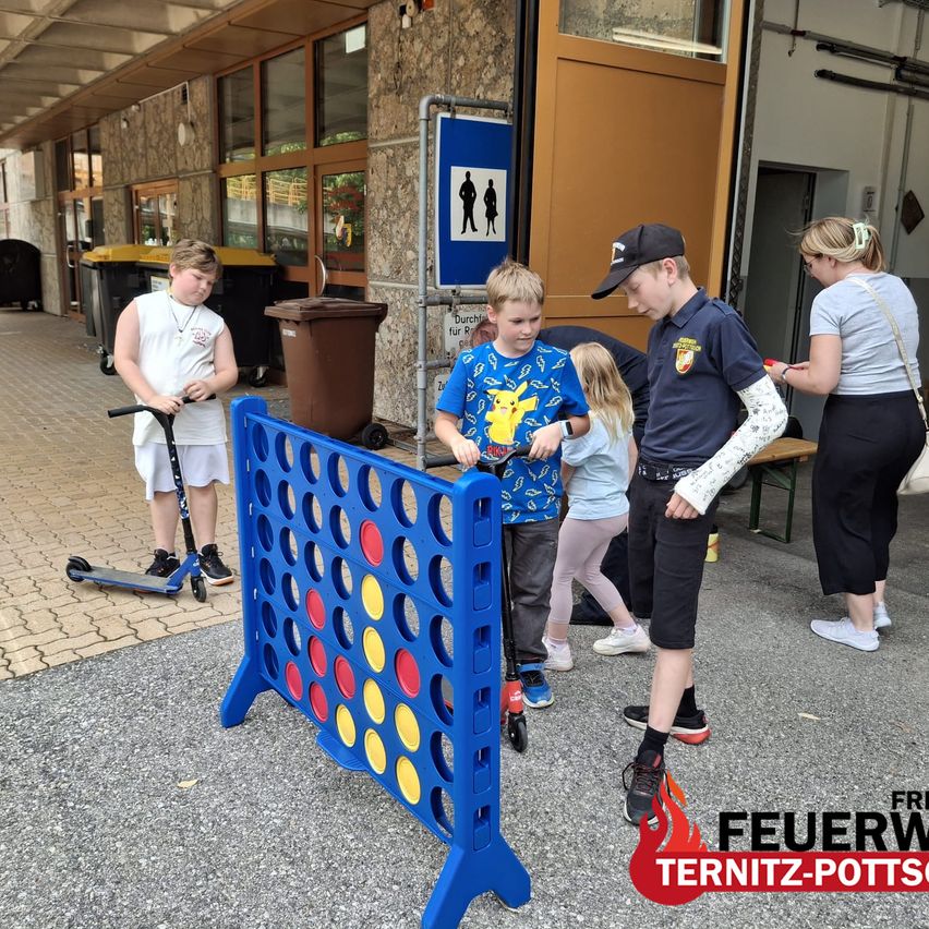 A group of children and adults are playing a game of connect four on a blue board outside. A boy in a Pikachu shirt is looking at the board.