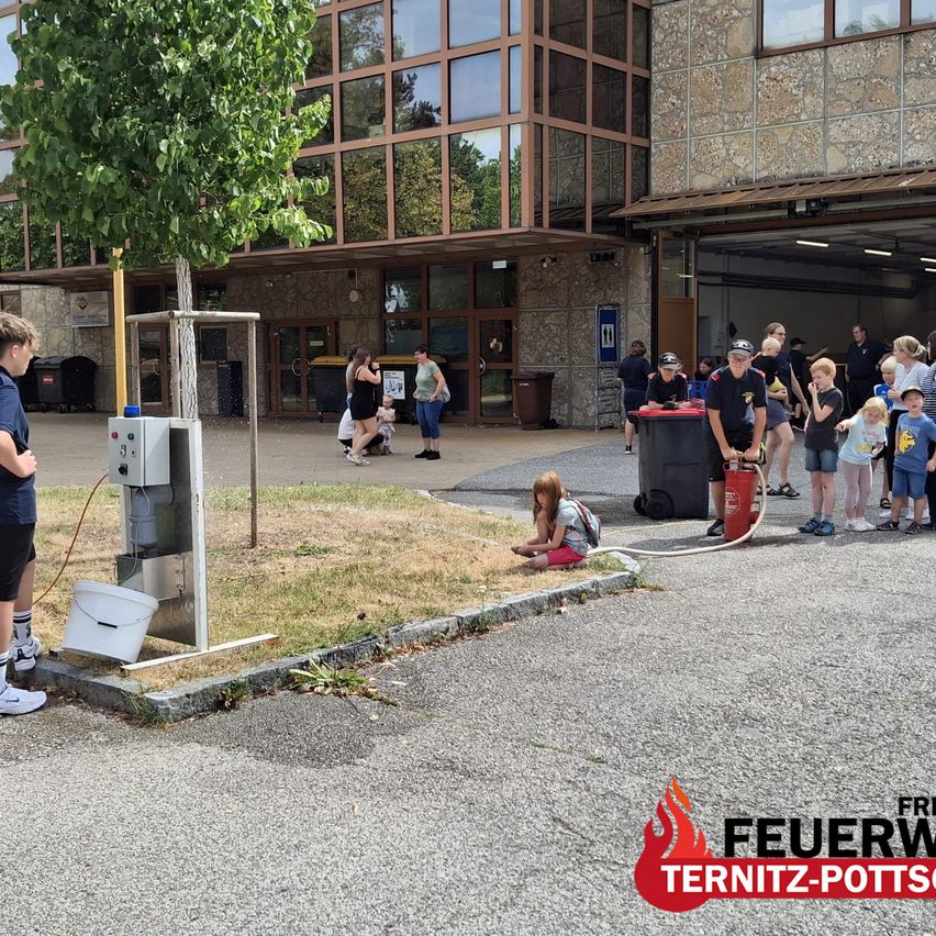 A group of people, including children, are gathered around a fire hydrant in front of a building. Some are observing while others are engaged in activities.