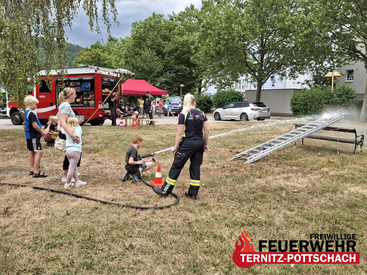 A firefighter is demonstrating how to use a water hose to a young boy while a woman and a girl watch. A fire truck is parked nearby with a ladder and tents.