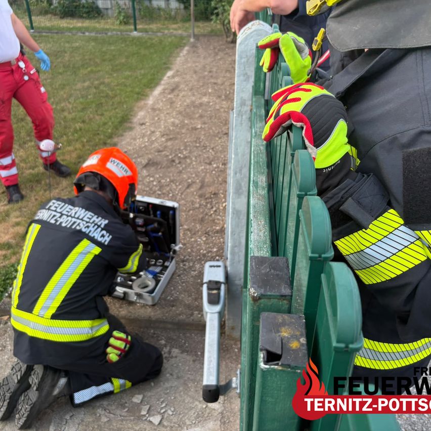 Ein Feuerwehrmann in einer reflektierenden Weste und Helm arbeitet an einem Metalltor, während ein anderer Feuerwehrmann hinter ihm steht. Die Szene spielt sich im Freien mit einem Grasbereich und einem Zaun im Hintergrund ab.